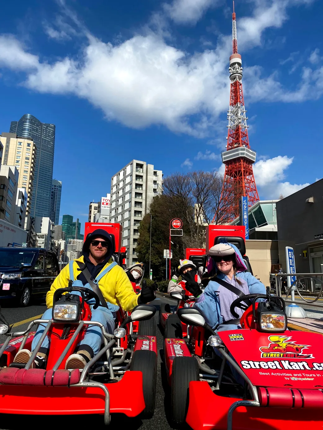 Group of people in red go-karts on a city street with Tokyo Tower in the background, smiling at the camera.