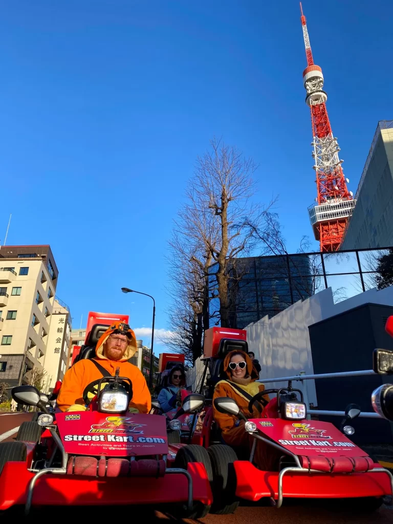 Two people in orange outfits ride red go-karts down a city street with Tokyo Tower in the background.