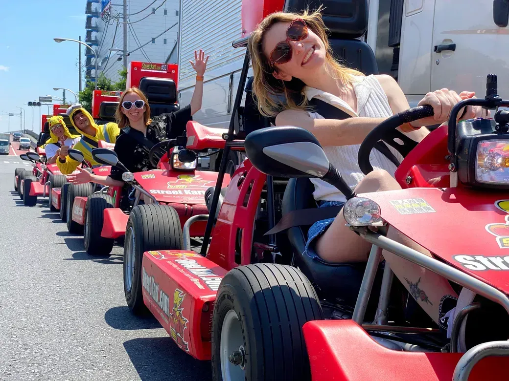 Smiling woman in sunglasses drives a red go-kart; a line of riders waves from behind on a sunny city street.