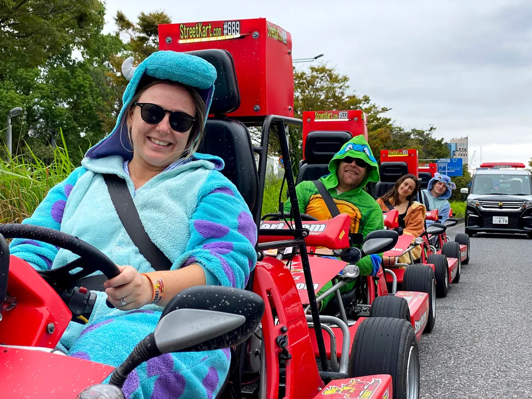Smiling woman in a blue koala onesie driving a red go-kart, wearing sunglasses, in a parade along a road.